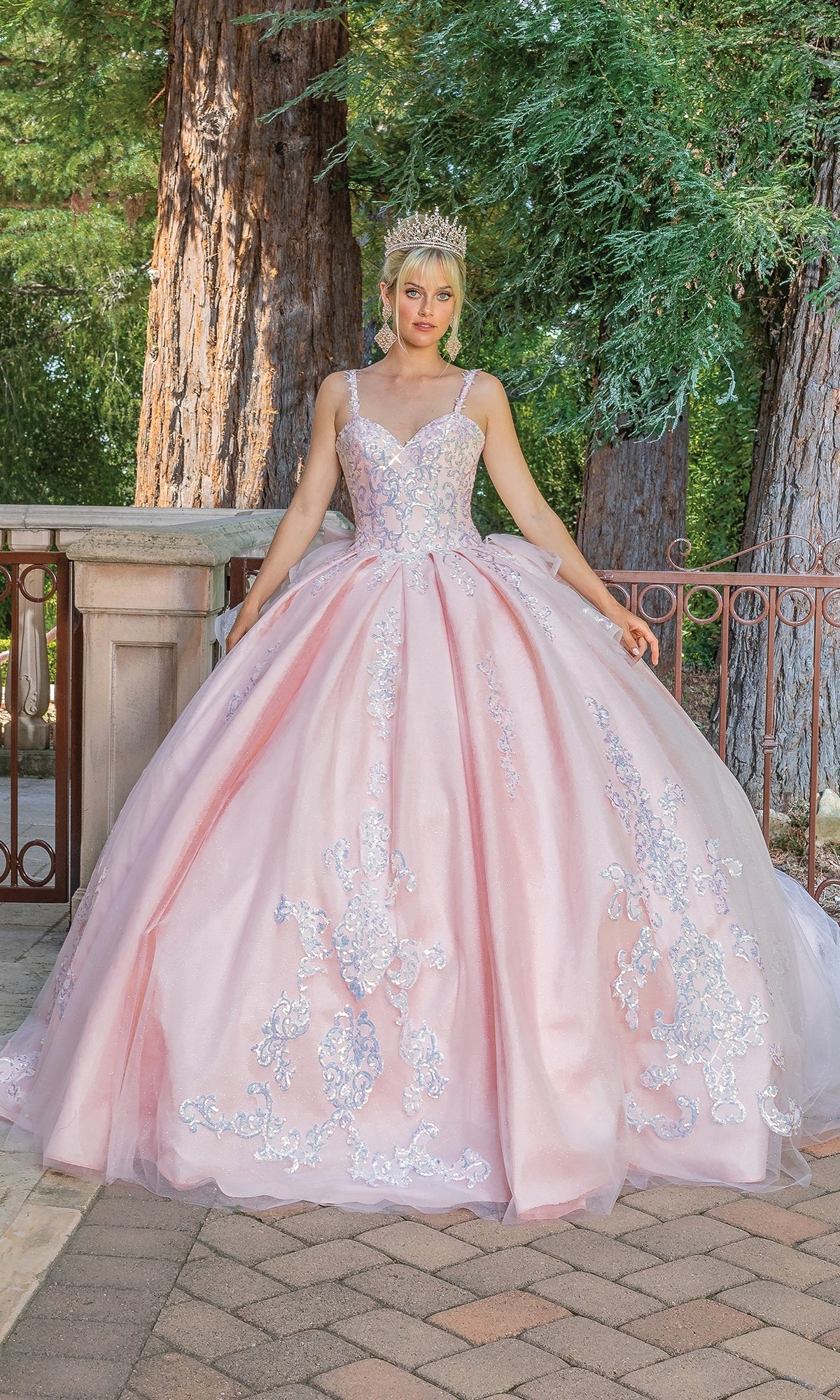 A young woman wearing the Quinceanera Dress 1652 by Dancing Queen, an a-line ball gown with a sweetheart neckline and floral embroidery, pairs it with a jeweled crown while standing outdoors by trees and a railing.
